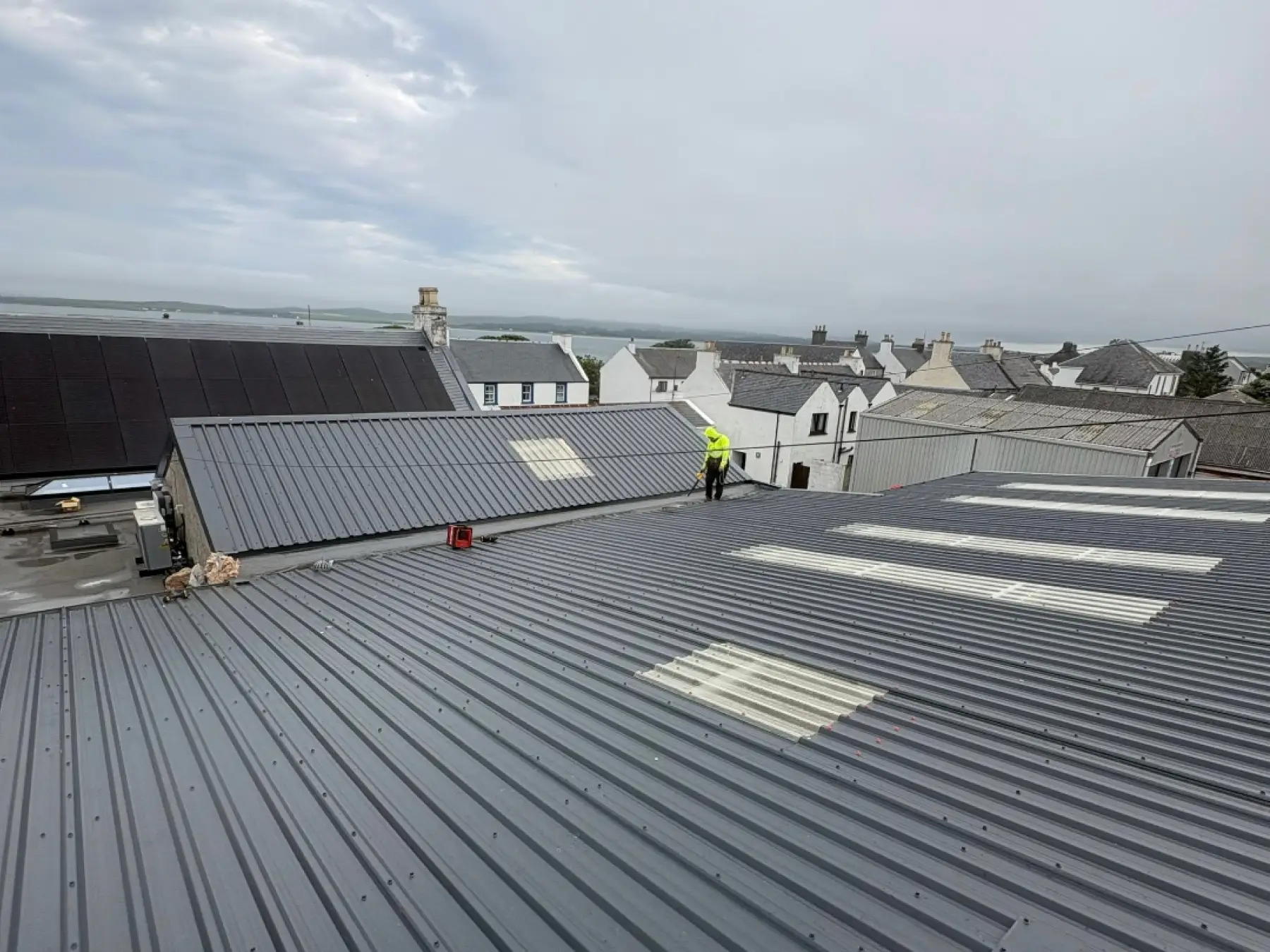 Roofer on new grey metal roofs with rooflights at Bowmore Hall, Islay, with village rooftops and water beyond