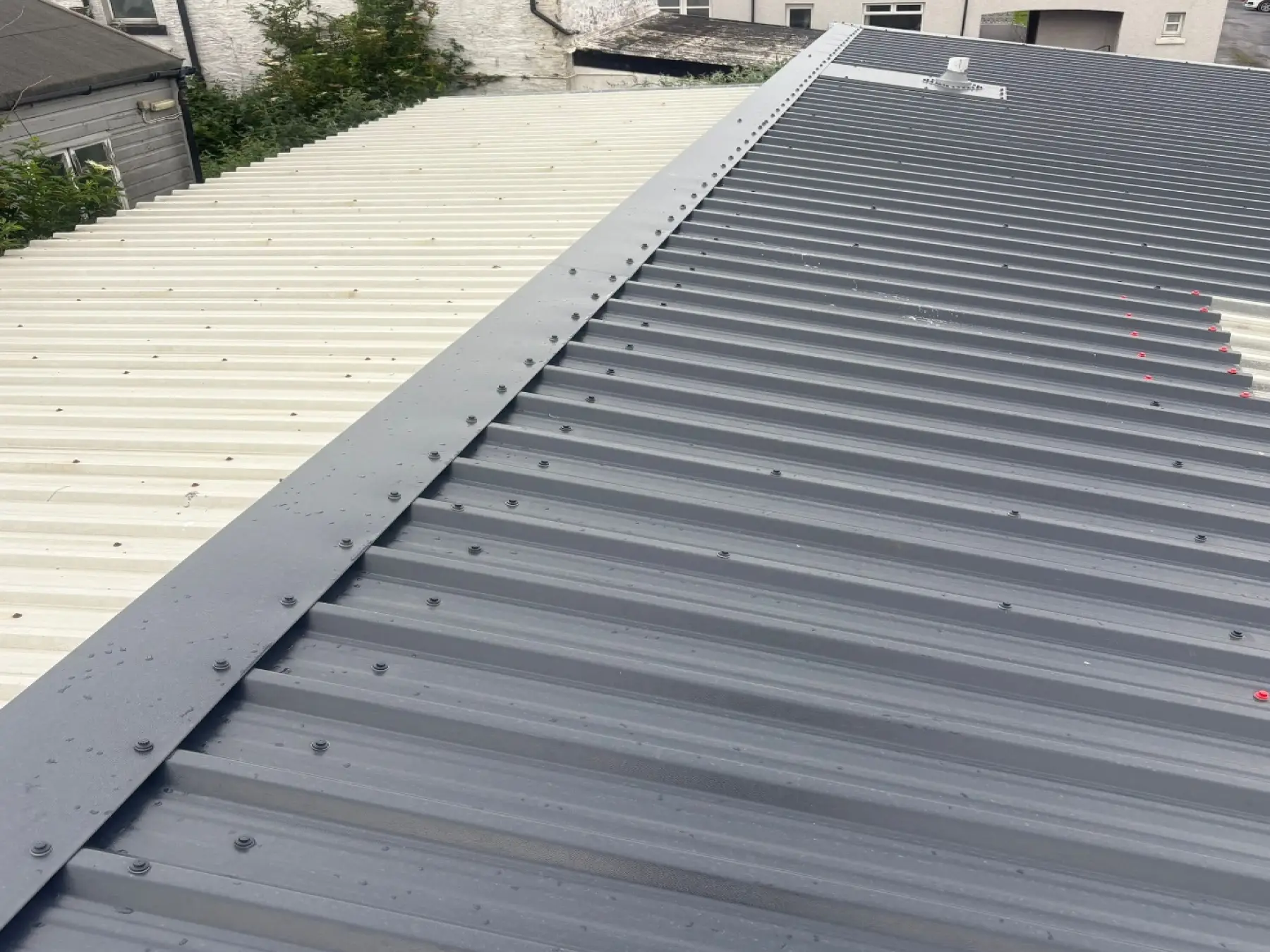 New dark grey profiled metal roof sheets and ridge flashing beside older cream sheets at Bowmore Hall, Islay