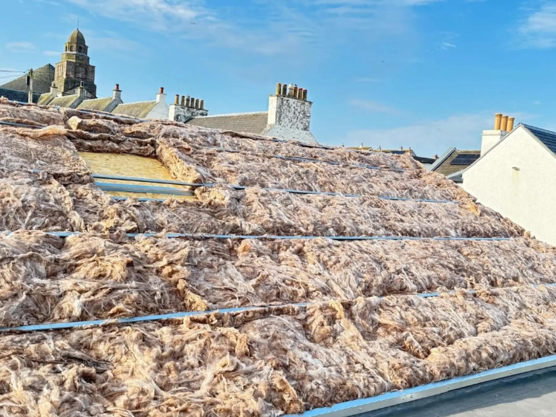 Mineral wool insulation laid between metal spacer rails on the pitched roof at Bowmore Hall, Islay, with chimneys beyond