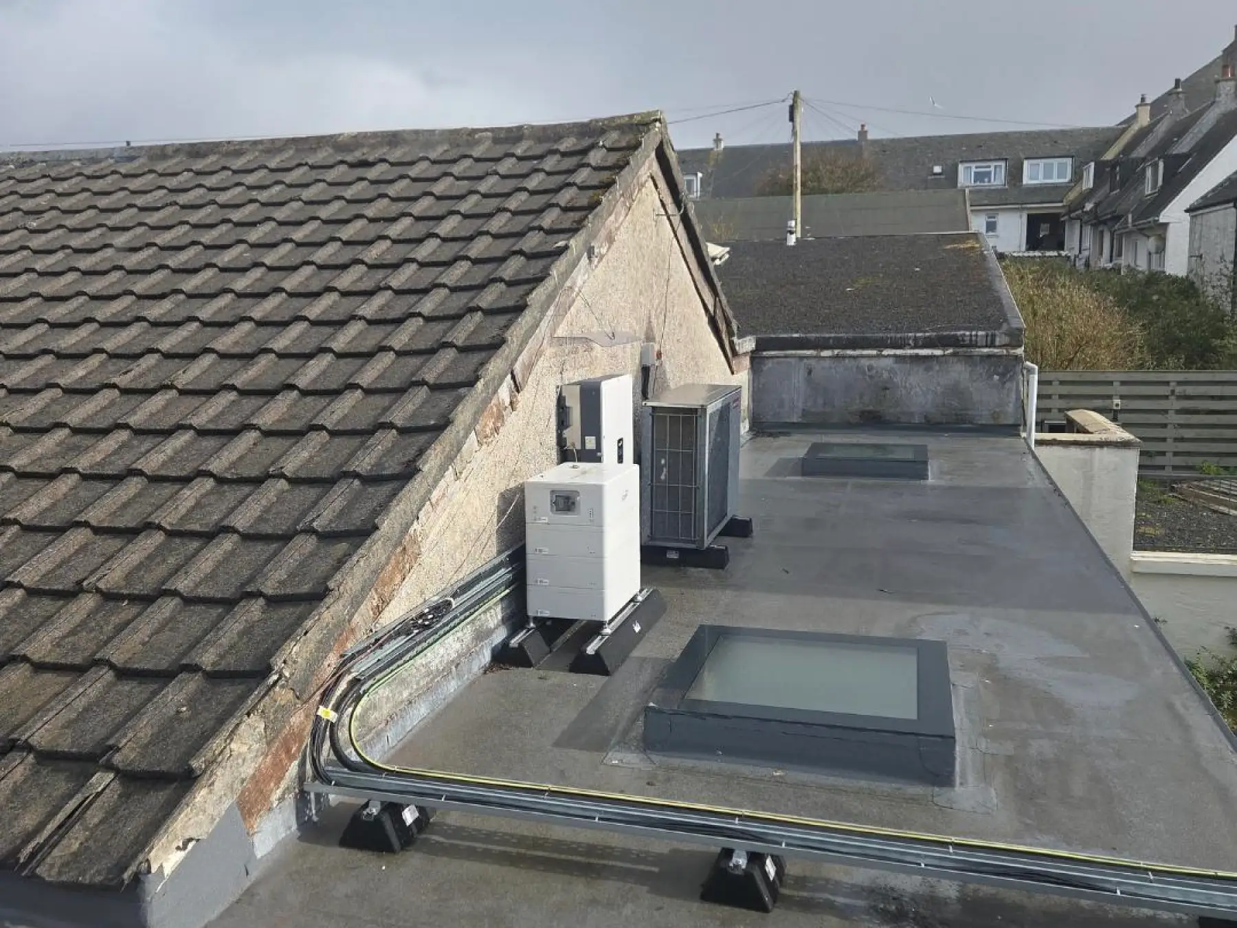 Grey membrane flat roof at Bowmore Hall, Islay, with glazed rooflights, rooftop plant and cable tray beside a tiled roof
