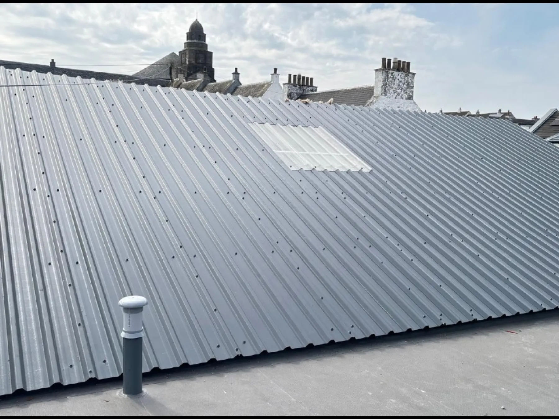 Grey profiled metal roof with a clear rooflight panel at Bowmore Hall, Islay, with chimneys and a stone tower behind