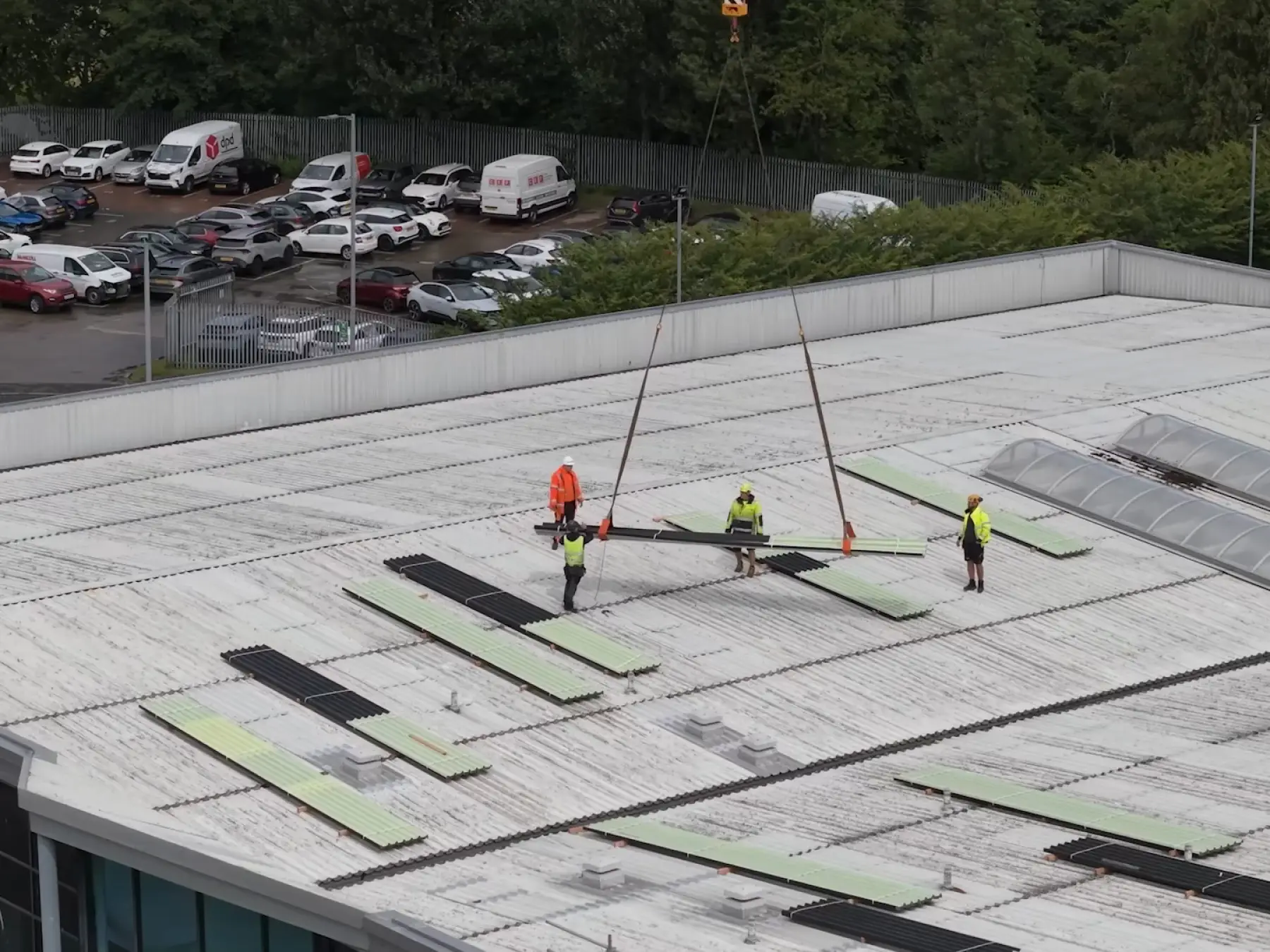 Aerial view of roofers in hi-vis guiding a crane-lifted bundle of profiled sheets onto an old corrugated roof