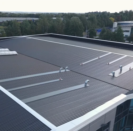 Dark grey profiled metal roof with white edge trims, rooflights and vent outlets, seen from above with trees beyond