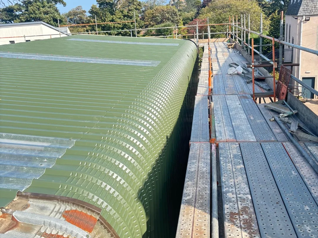 Curved green metal roof and rooflights beside scaffold access during school roofing work.