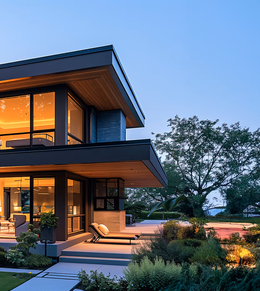 Modern two-story house with large glass windows illuminated warmly at dusk, surrounded by greenery and outdoor lounge chairs.