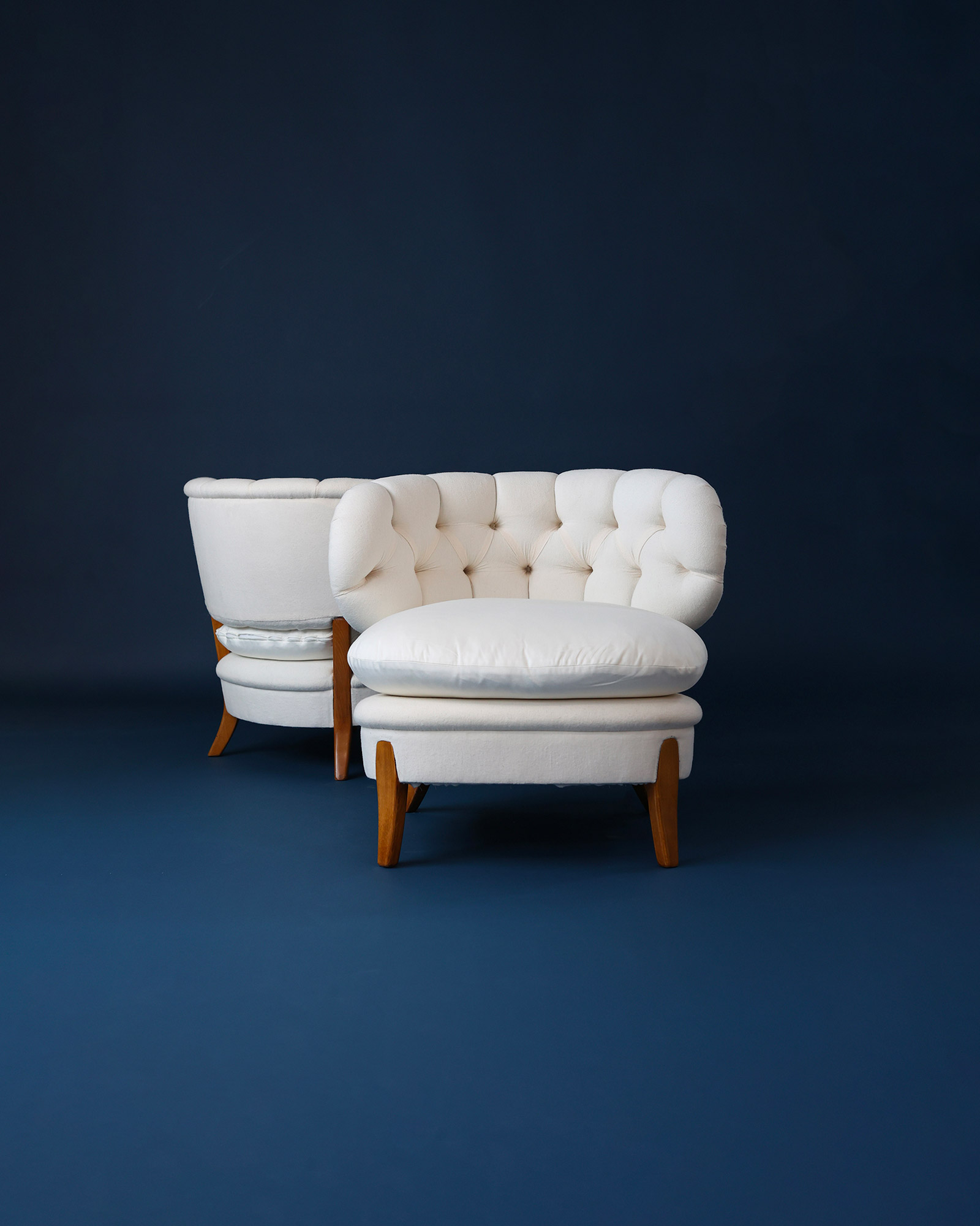 Another angled three-quarter perspective product photograph of two graceful white Otto Schulz upholstered easy chairs on a deep blue backdrop.