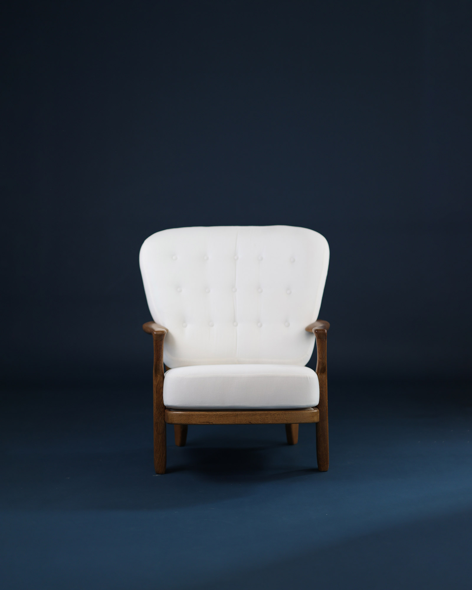 Juliette armchair by Guillerme et Chambron, front view showing the light wood frame with armrests, the high backrest with spindle structure, and the white upholstered seat against a dark blue background.