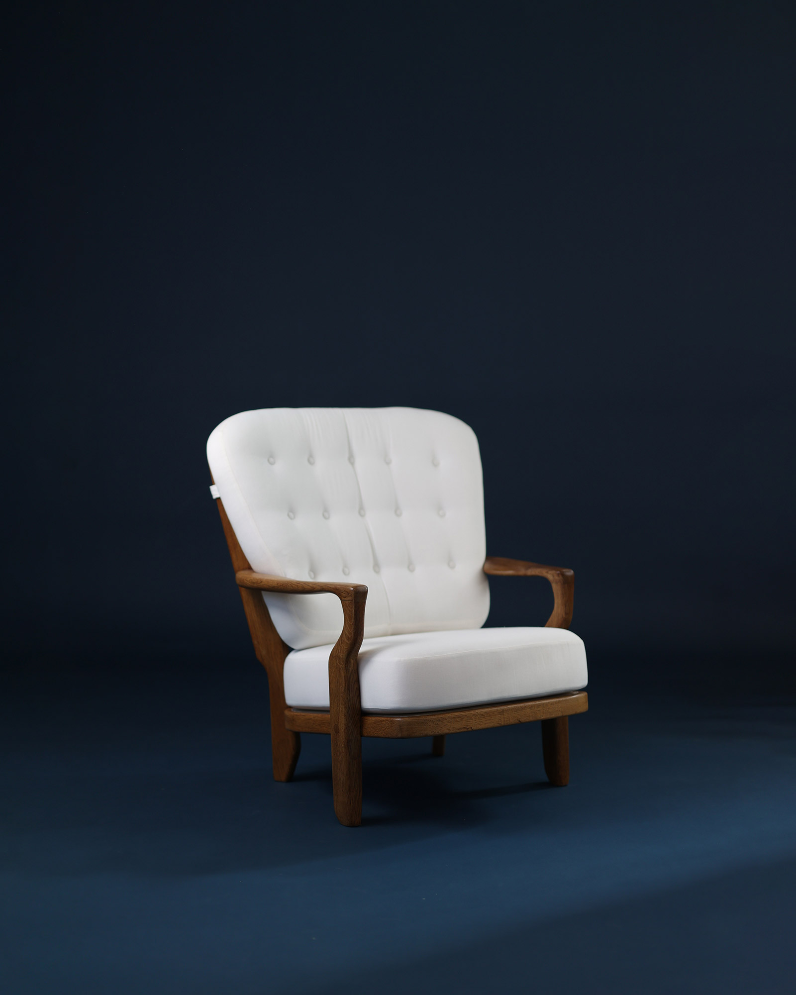 An angled three-quarter front view of the Juliette armchair by Guillerme et Chambron, displaying the profile of the light wood armrests and frame with the depth of the white seat against a dark blue background.