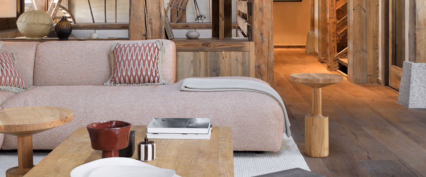 Salon avec canapé beige, coussins rouges, tables en bois, livres et décoration rustique en arrière-plan, La Ferme des Drus, Chamonix-Mont-Blanc, France.
