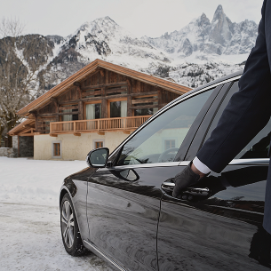 Person wearing gloves opening a black car door with a snowy mountain chalet in the background, La Ferme des Drus, Chamonix-Mont-Blanc, France.
