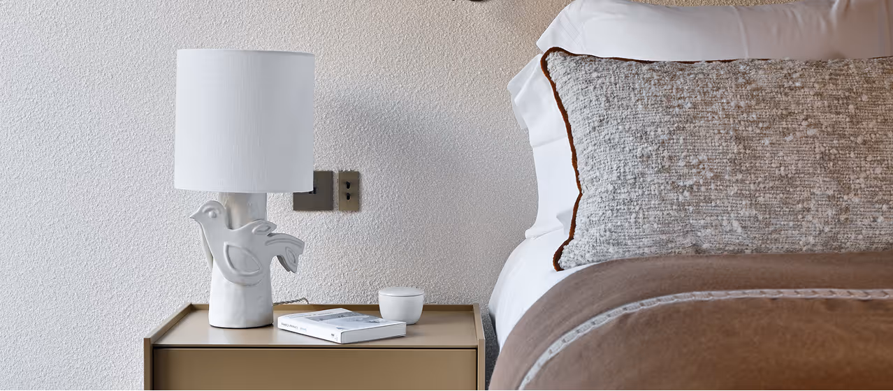 Lampe blanche avec base oiseau sur une table de chevet à côté d'un lit avec coussin beige, La Ferme des Drus, Chamonix-Mont-Blanc, France.