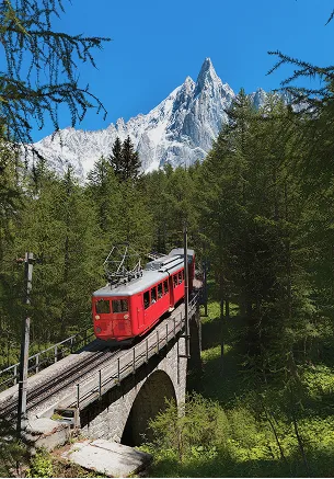 Un train rouge traverse un pont dans une forêt avec une montagne enneigée en arrière-plan, La Ferme des Drus, Chamonix-Mont-Blanc, France.
