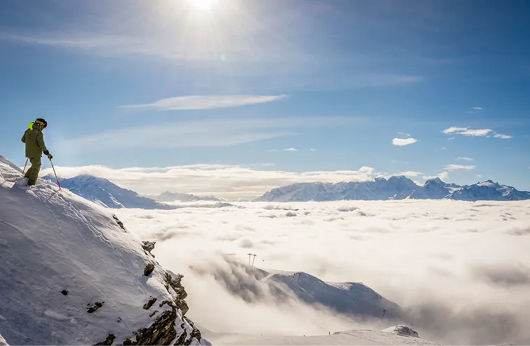 Un skieur vêtu de vert descend une pente enneigée avec un ciel bleu et une mer de nuages en arrière-plan, La Ferme des Drus, Chamonix-Mont-Blanc, France.