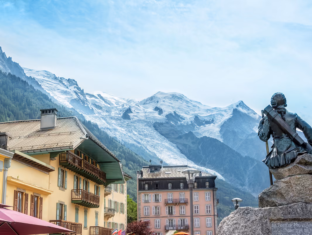 Statue sur la place du village avec des bâtiments de style alpin et une chaîne de montagnes enneigée en arrière-plan, La Ferme des Drus, Chamonix-Mont-Blanc, France.
