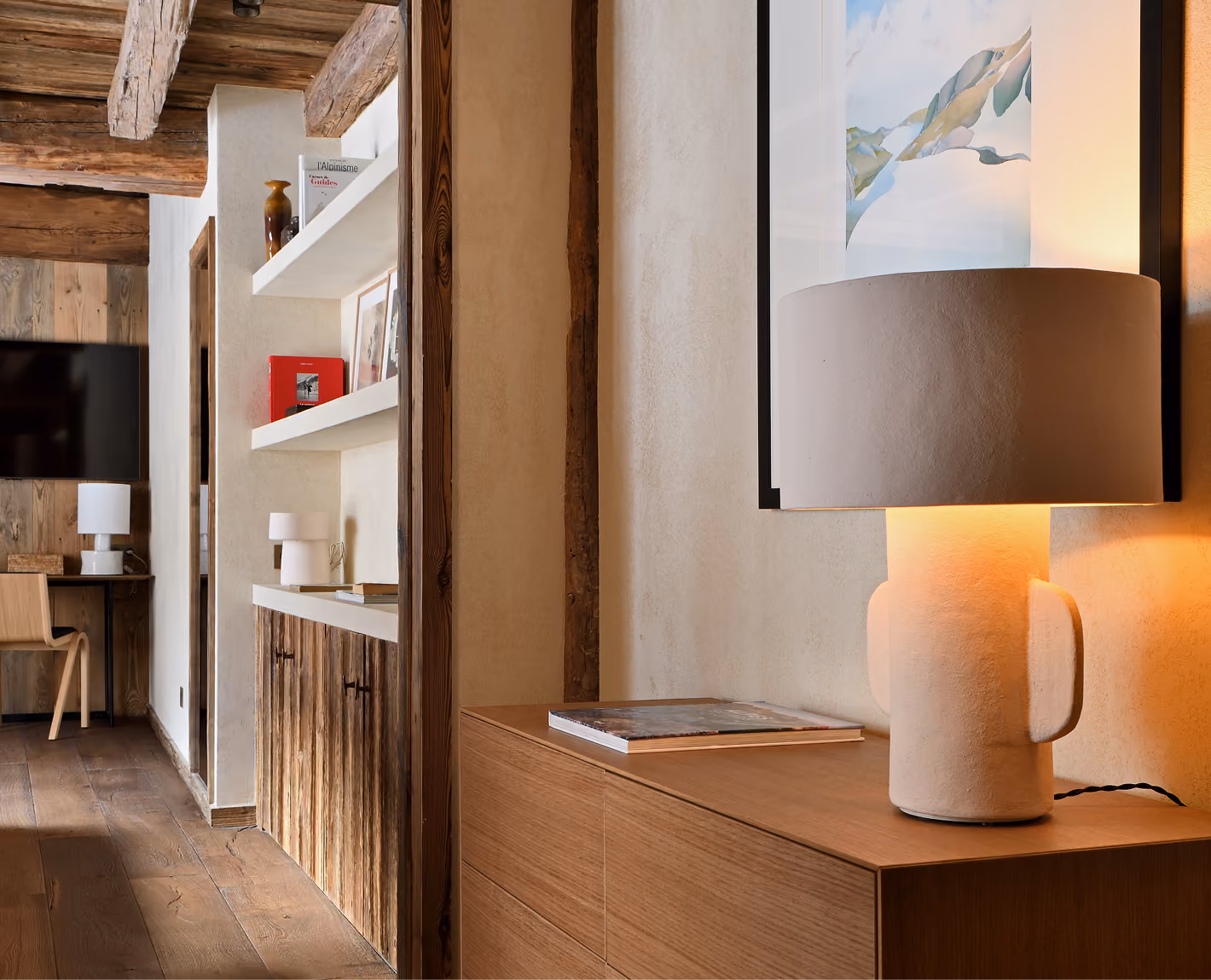 Lampe beige allumée sur meuble en bois avec peinture murale et étagères décorées en arrière-plan, La Ferme des Drus, Chamonix-Mont-Blanc, France.