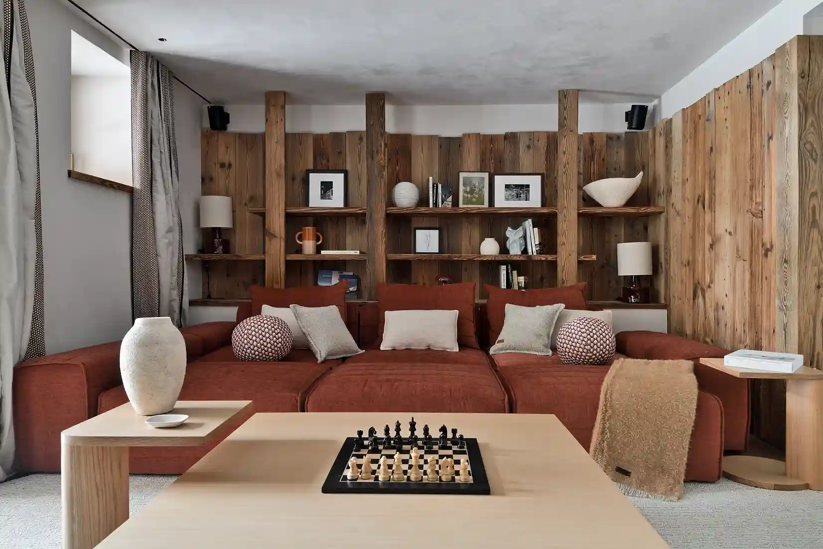 Salon moderne avec canapé rouge, coussins, table basse avec échiquier et étagères en bois au mur, La Ferme des Drus, Chamonix-Mont-Blanc, France.