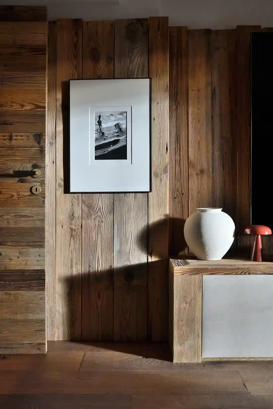 Mur en bois avec cadre photo noir et blanc, vase blanc et lampe rouge sur meuble bas en bois, La Ferme des Drus, Chamonix-Mont-Blanc, France.