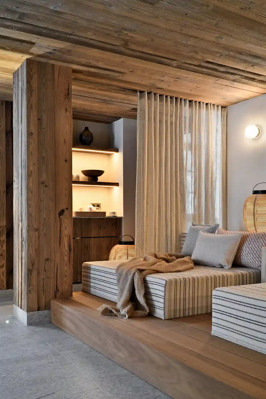 Coin salon avec banquette rayée, coussins, plaid, bois naturel et lumière douce derrière un rideau, La Ferme des Drus, Chamonix-Mont-Blanc, France.
