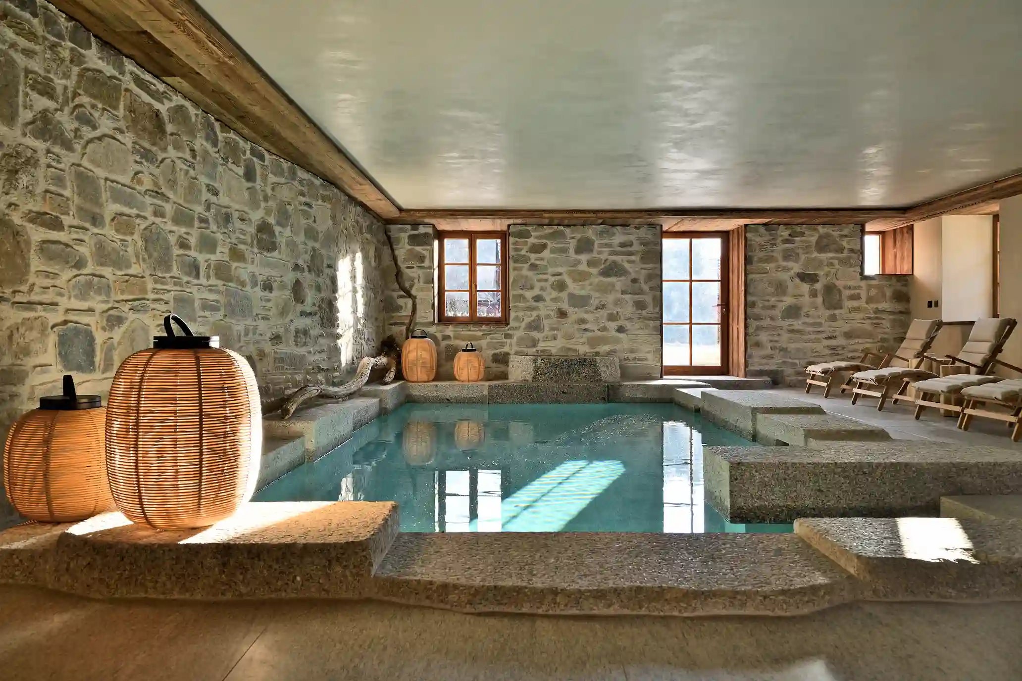 Piscine intérieure avec murs en pierre, lanternes en osier et chaises longues en bois avec coussins, La Ferme des Drus, Chamonix-Mont-Blanc, France.