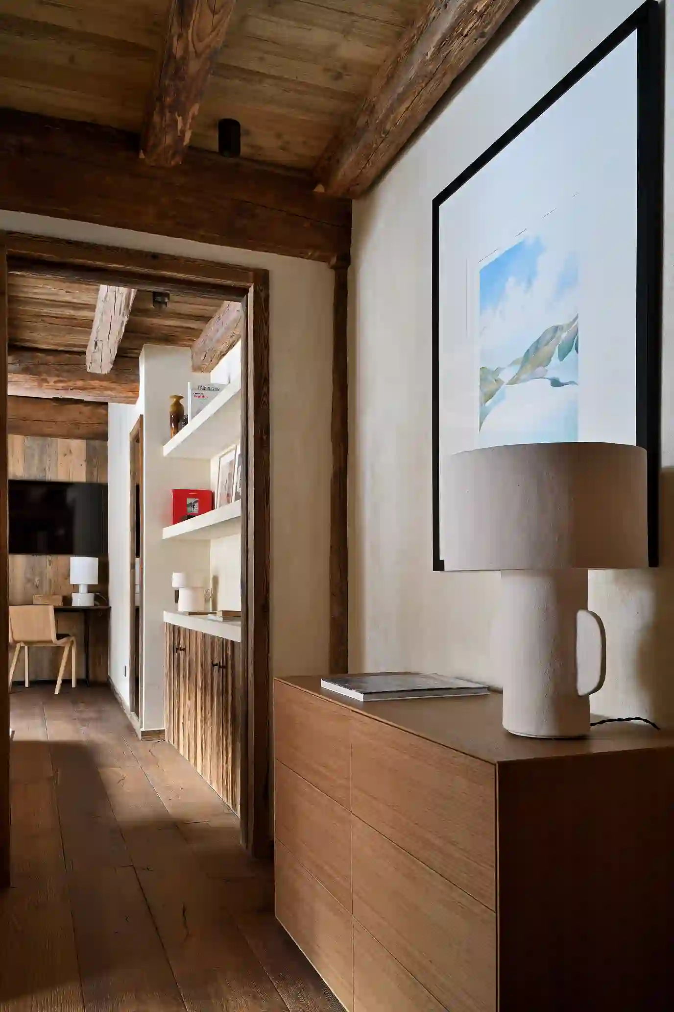 Intérieur en bois chaleureux avec commode, lampe beige et tableau montagne, couloir menant à un bureau, La Ferme des Drus, Chamonix-Mont-Blanc, France.