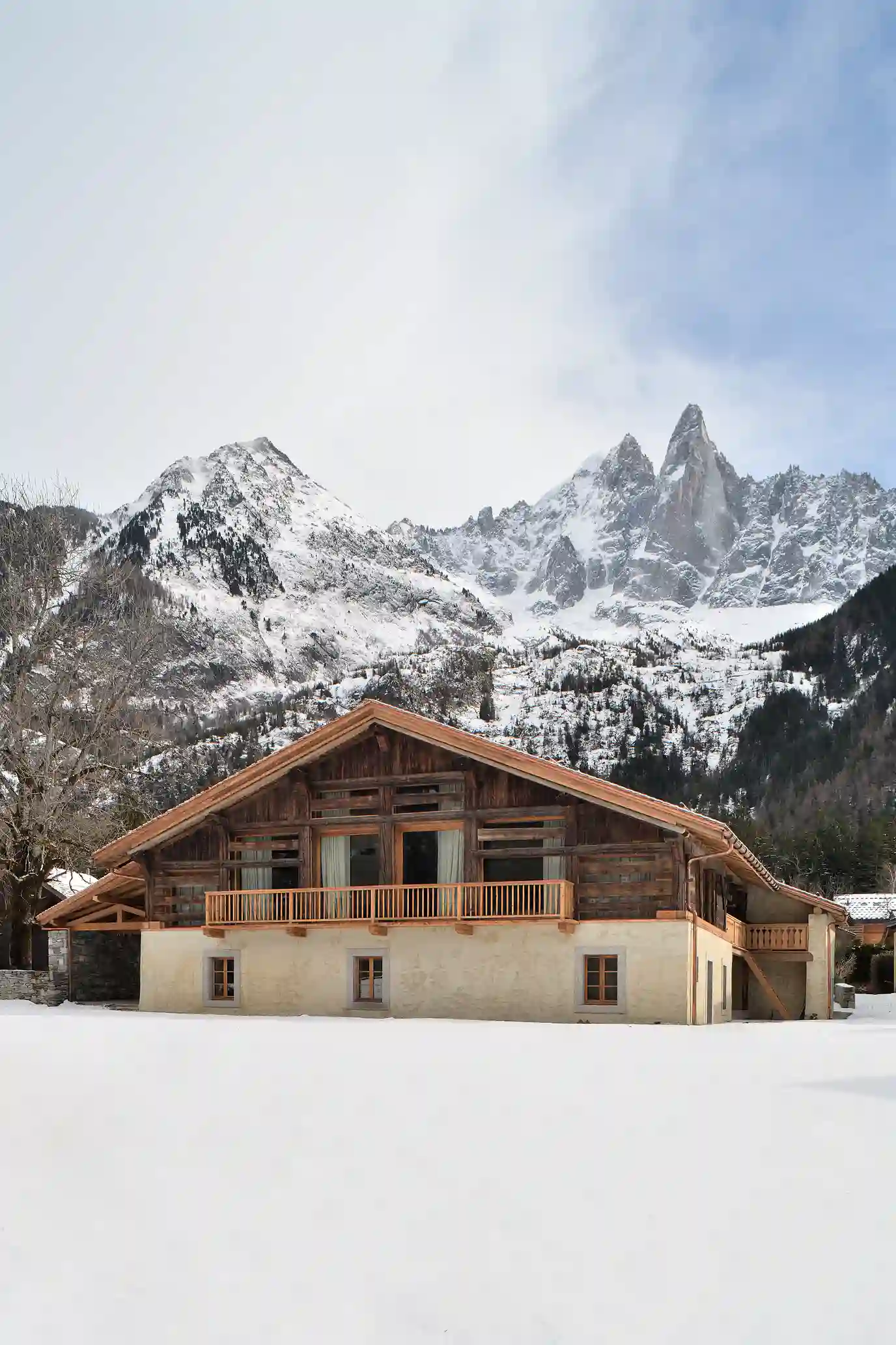 Chalet en bois et pierre devant un paysage de montagnes enneigées sous un ciel clair, La Ferme des Drus, Chamonix-Mont-Blanc, France.