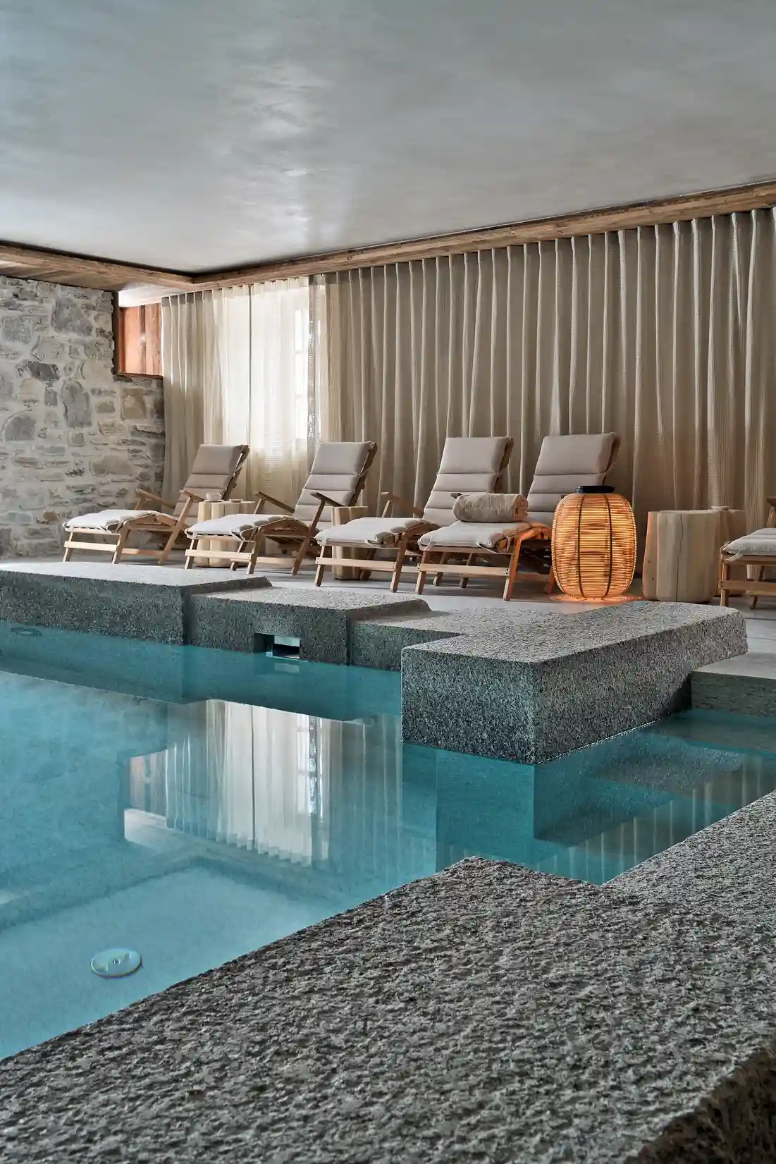 Piscine intérieure avec chaises longues en bois et lampe posée près d’un mur en pierre, La Ferme des Drus, Chamonix-Mont-Blanc, France.