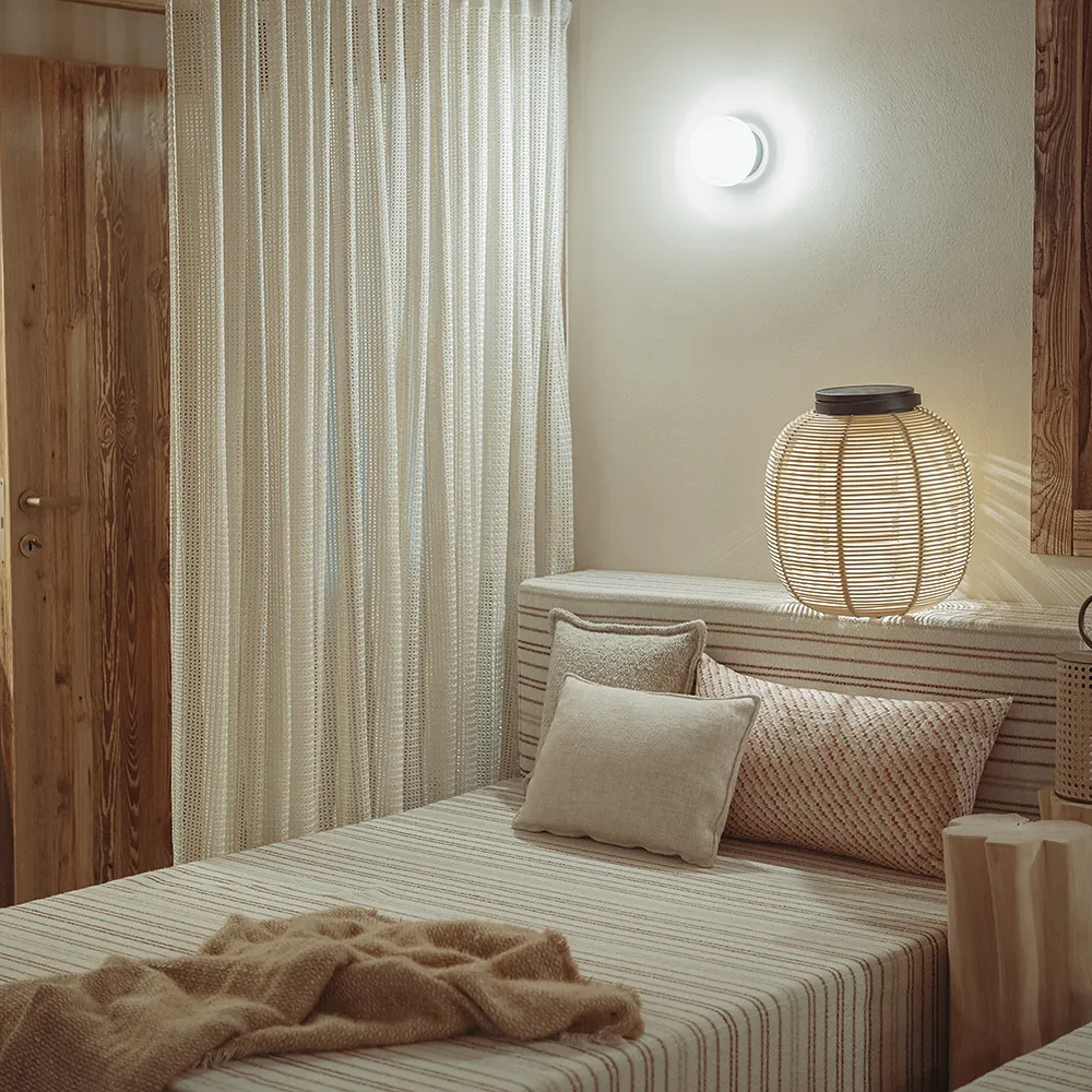 Chambre beige avec lit rayé, coussins, plaid et lampe en rotin éclairée sur le mur, La Ferme des Drus, Chamonix-Mont-Blanc, France.