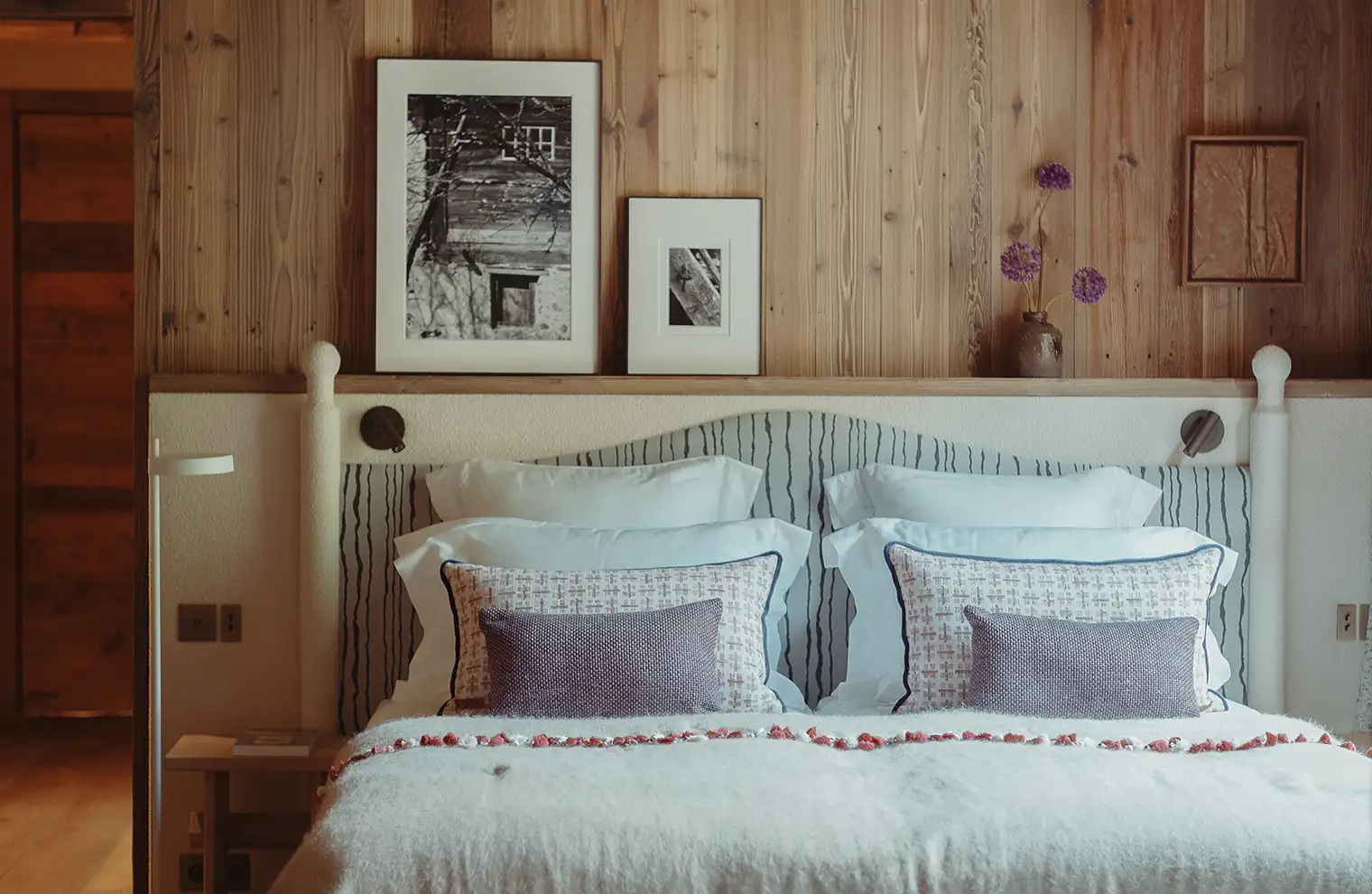 Chambre avec lit double, coussins décoratifs et murs en bois avec cadres photo et vase fleuri, La Ferme des Drus, Chamonix-Mont-Blanc, France.