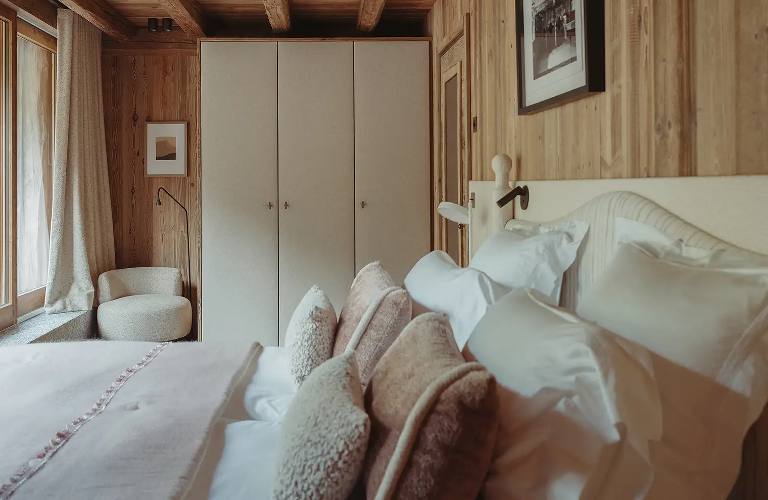 Chambre en bois avec lit décoré de coussins beiges, fauteuil près d'une fenêtre et armoire claire, La Ferme des Drus, Chamonix-Mont-Blanc, France.