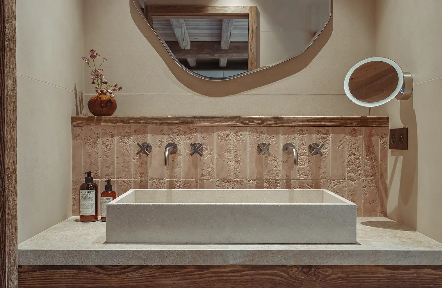 Lave-mains en pierre beige avec robinet mural, miroir irrégulier, et flacon de savon liquide brun, La Ferme des Drus, Chamonix-Mont-Blanc, France.