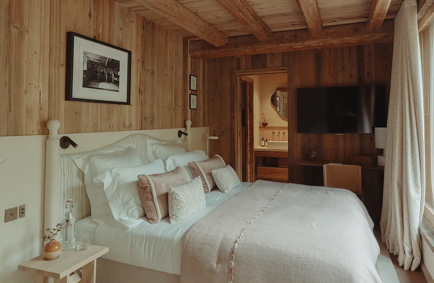 Chambre à coucher avec murs en bois, lit double blanc, coussins, télé et salle de bain attenante, La Ferme des Drus, Chamonix-Mont-Blanc, France.