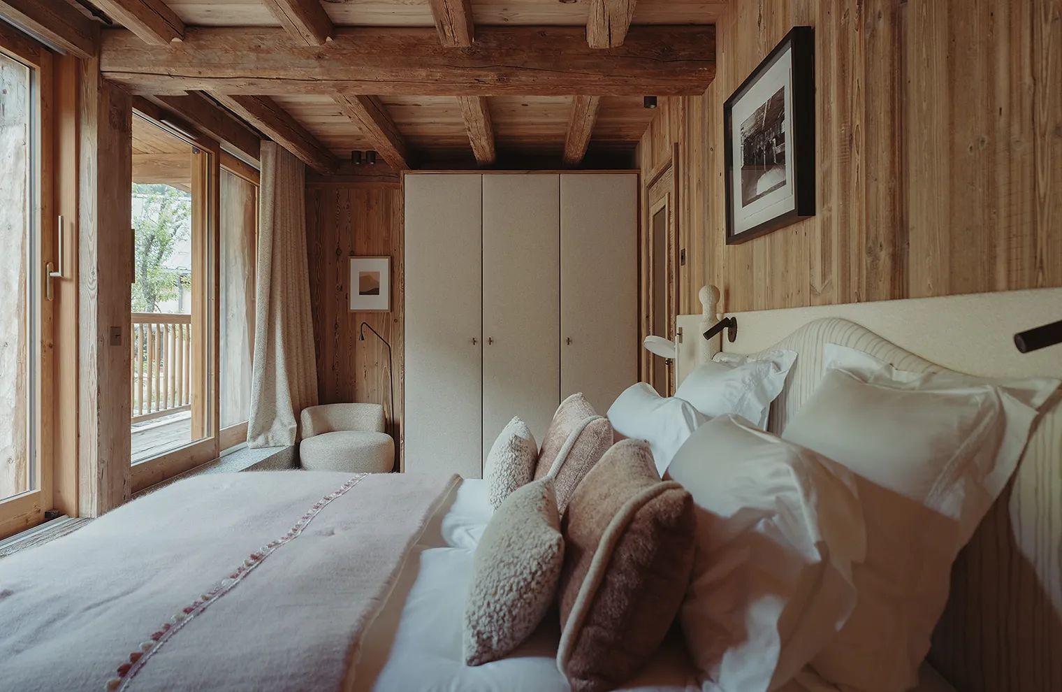 Chambre en bois avec lit double, coussins, chaise, armoire et balcon extérieur visible, La Ferme des Drus, Chamonix-Mont-Blanc, France.