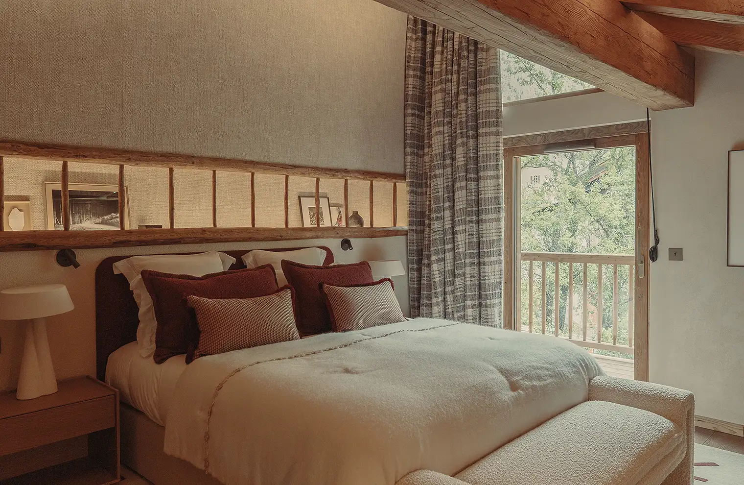 Chambre avec lit double, coussins bordeaux, lampes blanches, et terrasse bois avec vue sur arbres, La Ferme des Drus, Chamonix-Mont-Blanc, France.