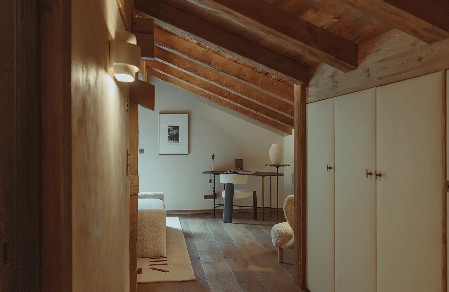 Bureau avec chaise blanche sous plafond incliné en bois dans une pièce chaleureuse avec canapé, La Ferme des Drus, Chamonix-Mont-Blanc, France.