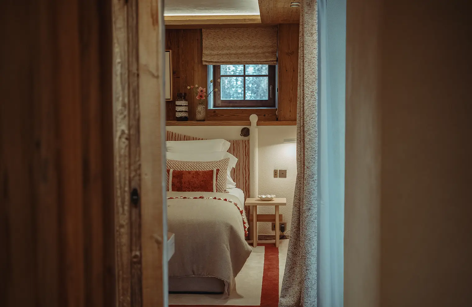 Chambre cosy avec lit beige, coussins décoratifs et fenêtre avec volet en bois, La Ferme des Drus, Chamonix-Mont-Blanc, France.