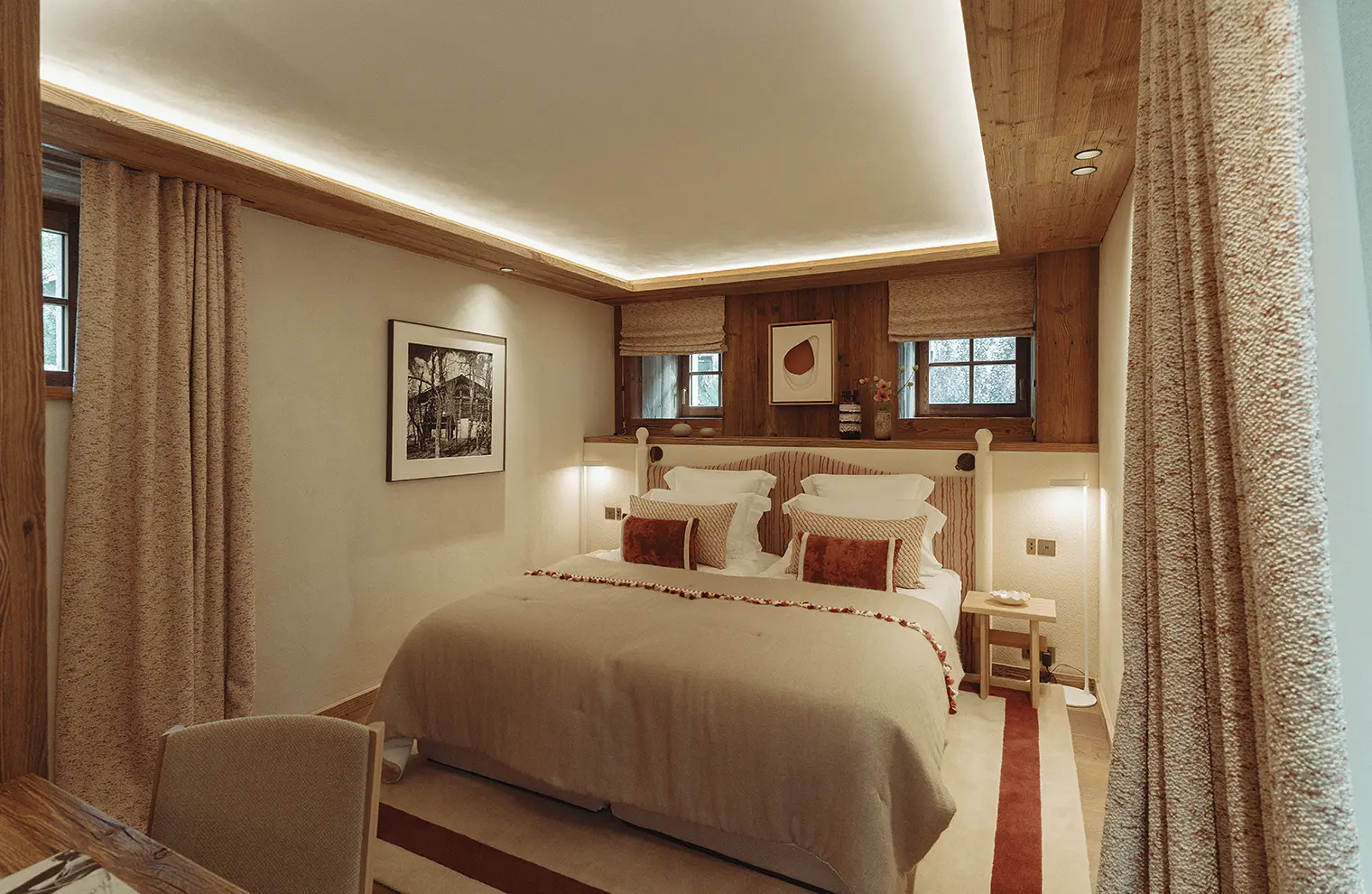 Chambre cosy avec grand lit double, draps beige, coussins rouges, murs bois et tentures épaisses, La Ferme des Drus, Chamonix-Mont-Blanc, France.