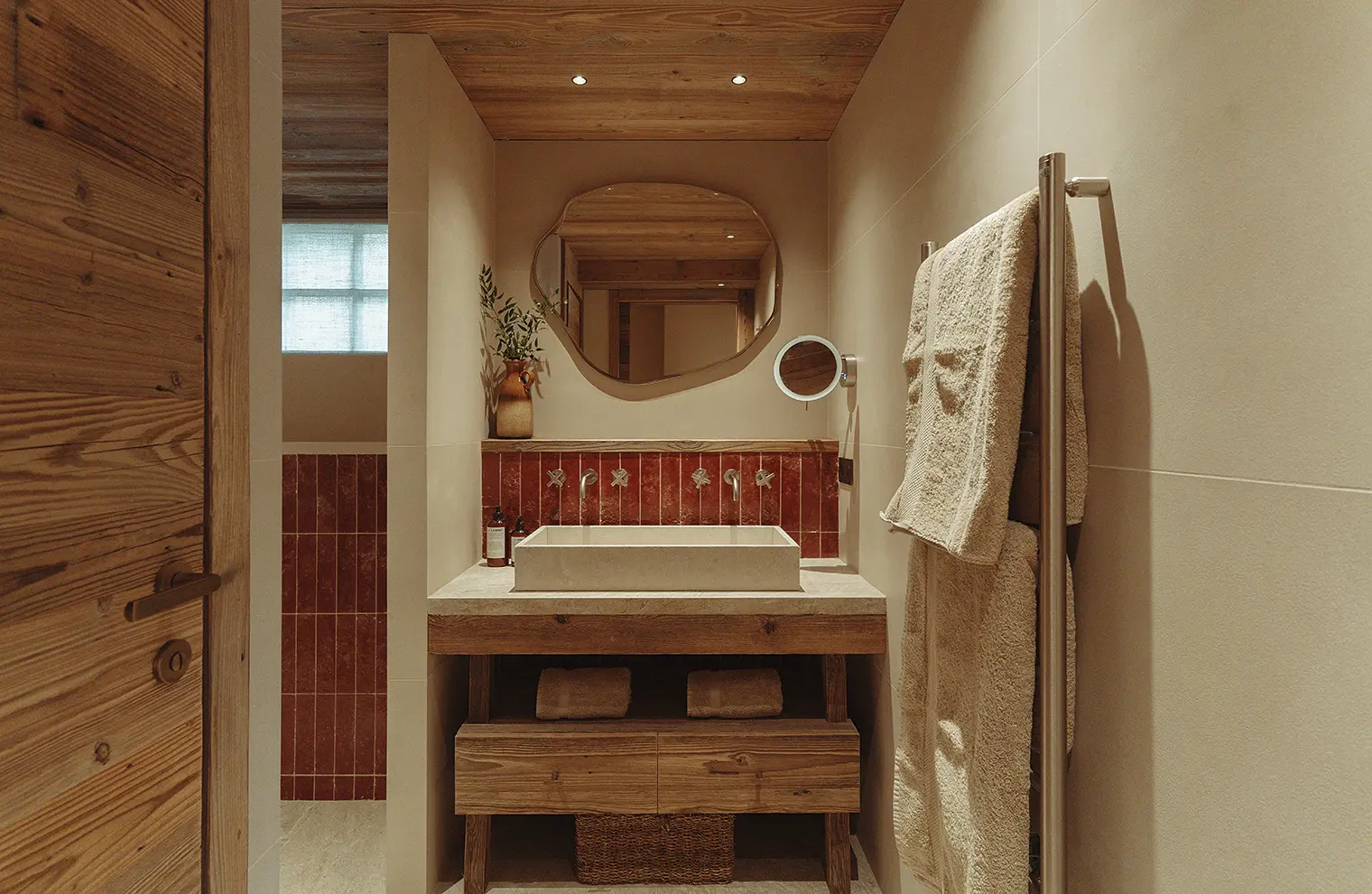 Salle de bain avec lavabo rectangulaire, miroir irrégulier, serviettes beiges et murs beige avec carrelage rouge, La Ferme des Drus, Chamonix-Mont-Blanc, France.