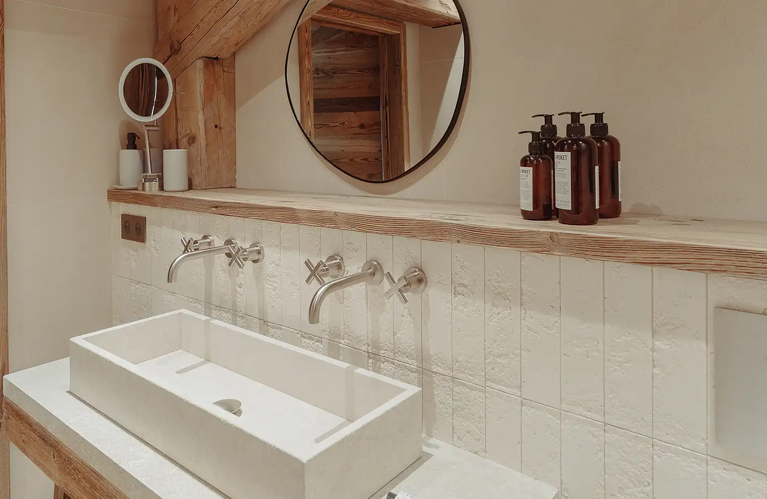 Lavabo rectangulaire en pierre avec robinetterie murale, miroir rond et flacons de savon dans une salle de bain en bois, La Ferme des Drus, Chamonix-Mont-Blanc, France.