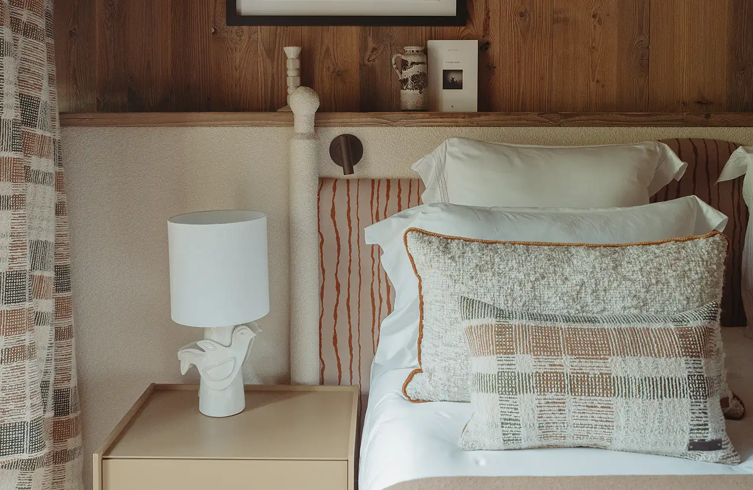 Coin chambre avec table de nuit beige, lampe blanche, coussins beiges et tête de lit à rayures, La Ferme des Drus, Chamonix-Mont-Blanc, France.