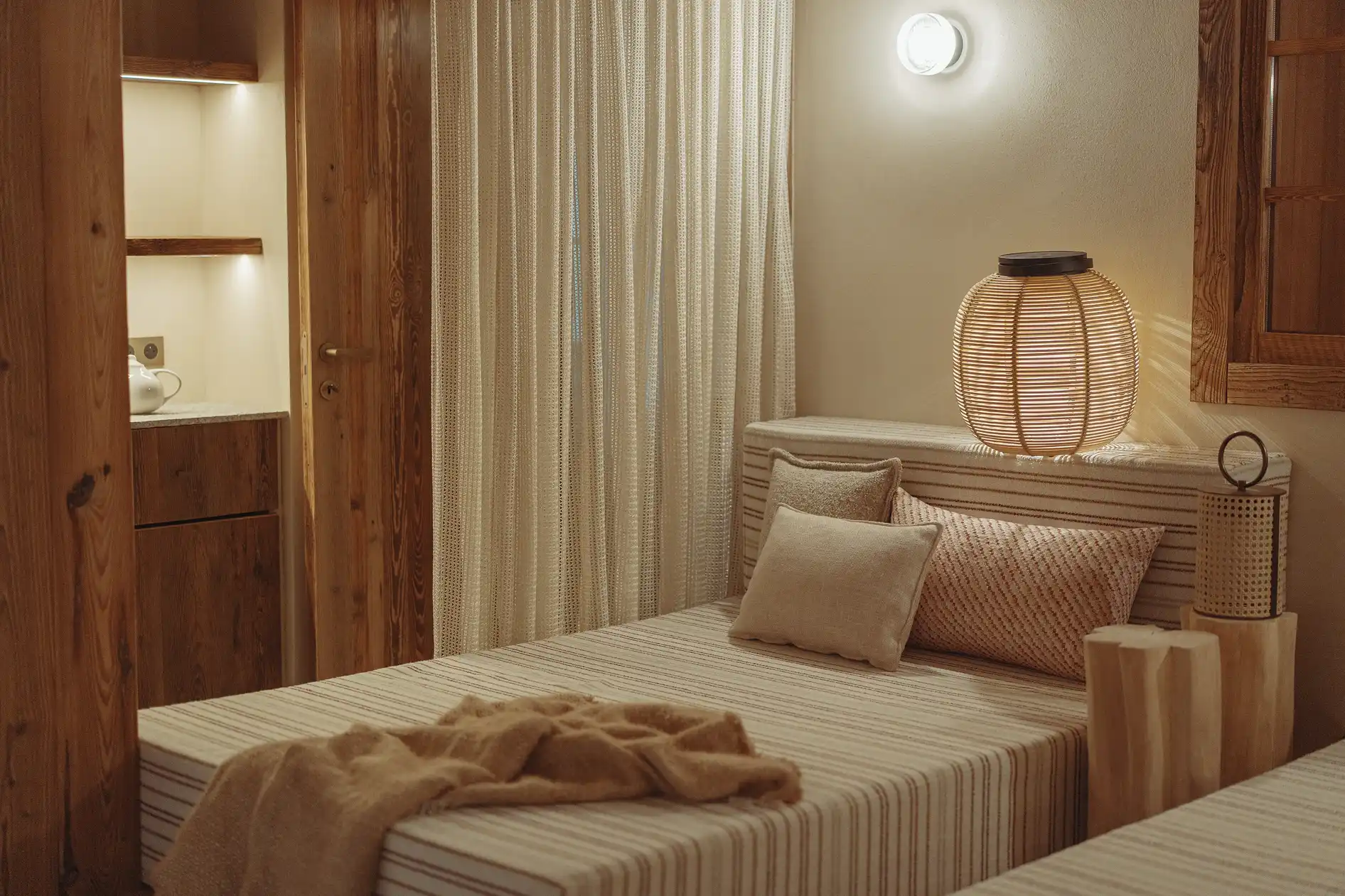 Chambre avec lit simple, coussins, lampe en osier et rideaux beiges dans un décor chaleureux en bois, La Ferme des Drus, Chamonix-Mont-Blanc, France.