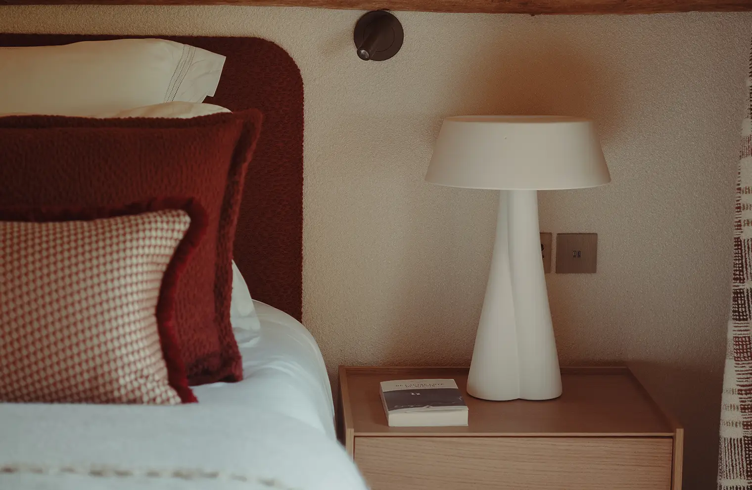 Table de chevet avec lampe blanche et livre à côté d'un lit avec coussins rouges, La Ferme des Drus, Chamonix-Mont-Blanc, France.
