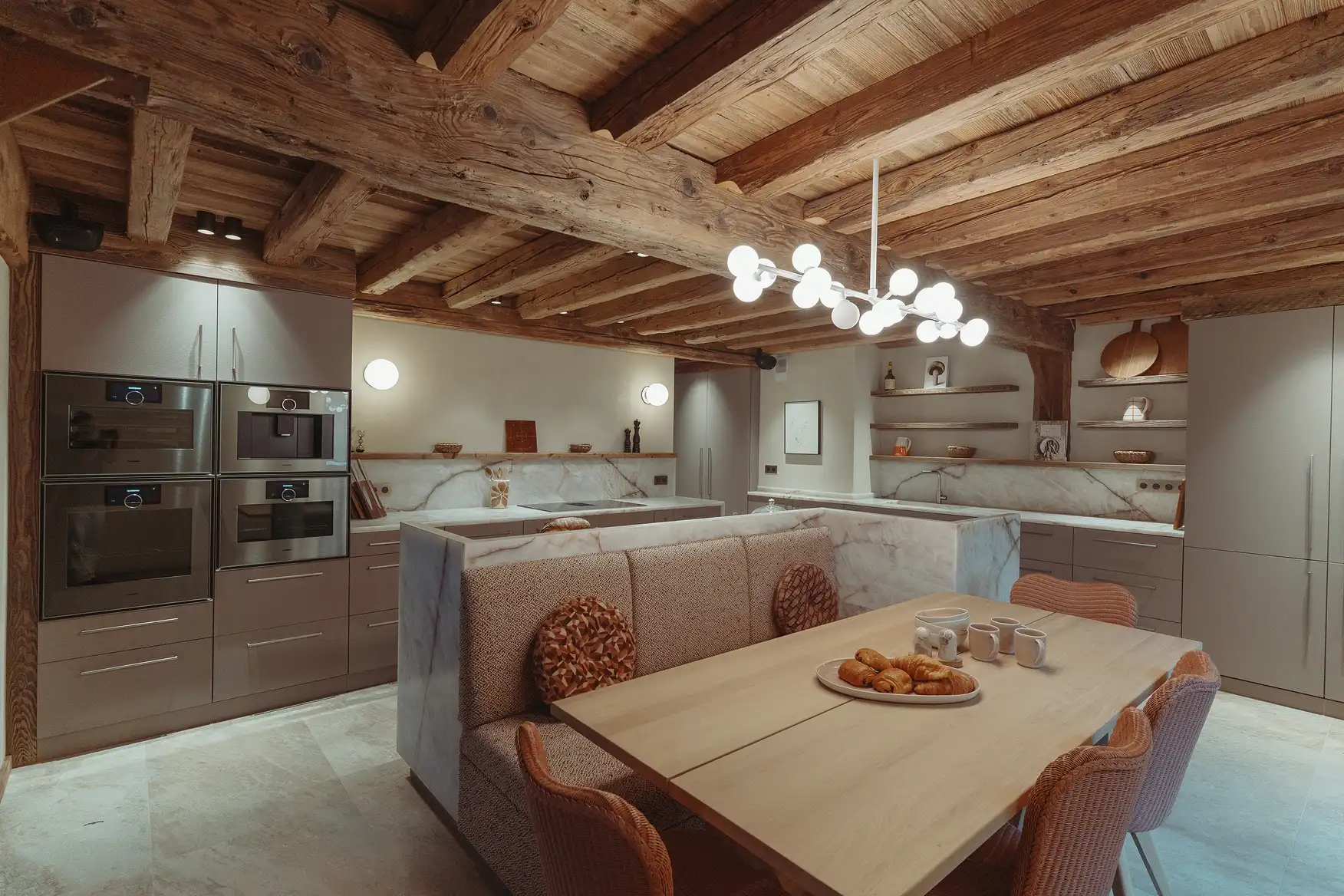 Cuisine moderne avec murs en bois, meubles gris, table en bois avec croissants et sièges en tissu, La Ferme des Drus, Chamonix-Mont-Blanc, France.