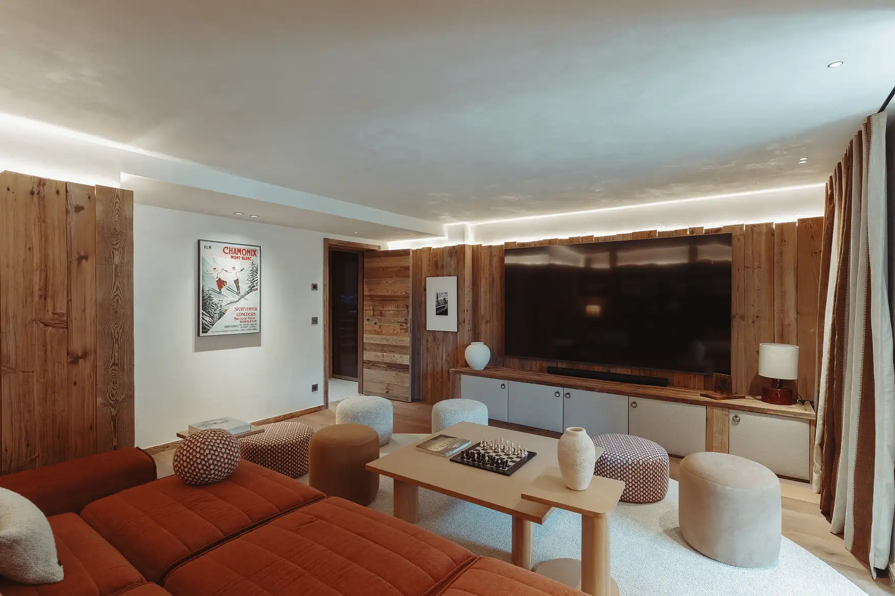 Salon moderne avec canapé orange, plusieurs poufs, table basse avec échiquier et grande télévision murale, La Ferme des Drus, Chamonix-Mont-Blanc, France.