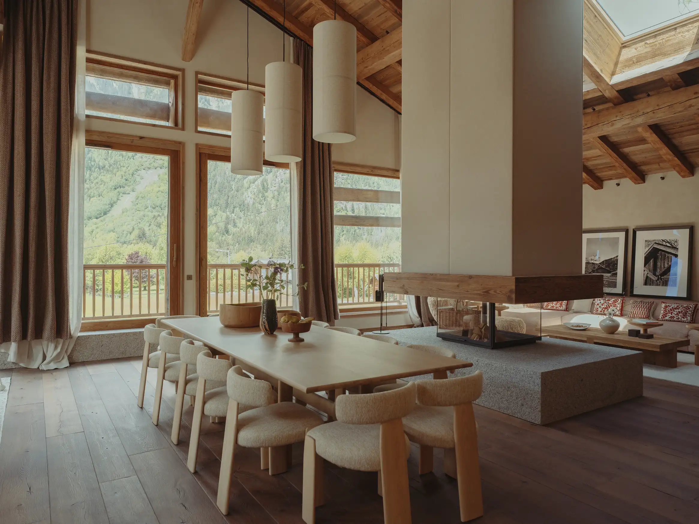 Salle à manger en bois avec grande table, chaises beige, cheminée centrale et grandes fenêtres sur paysage montagneux, La Ferme des Drus, Chamonix-Mont-Blanc, France.