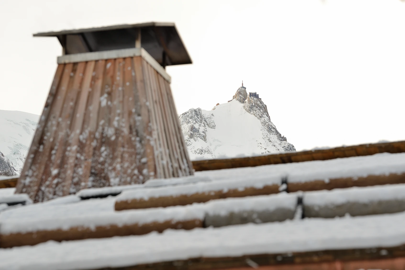 Cheminée en bois sur toit enneigé avec montagne enneigée et sommet rocheux en arrière-plan, La Ferme des Drus, Chamonix-Mont-Blanc, France.