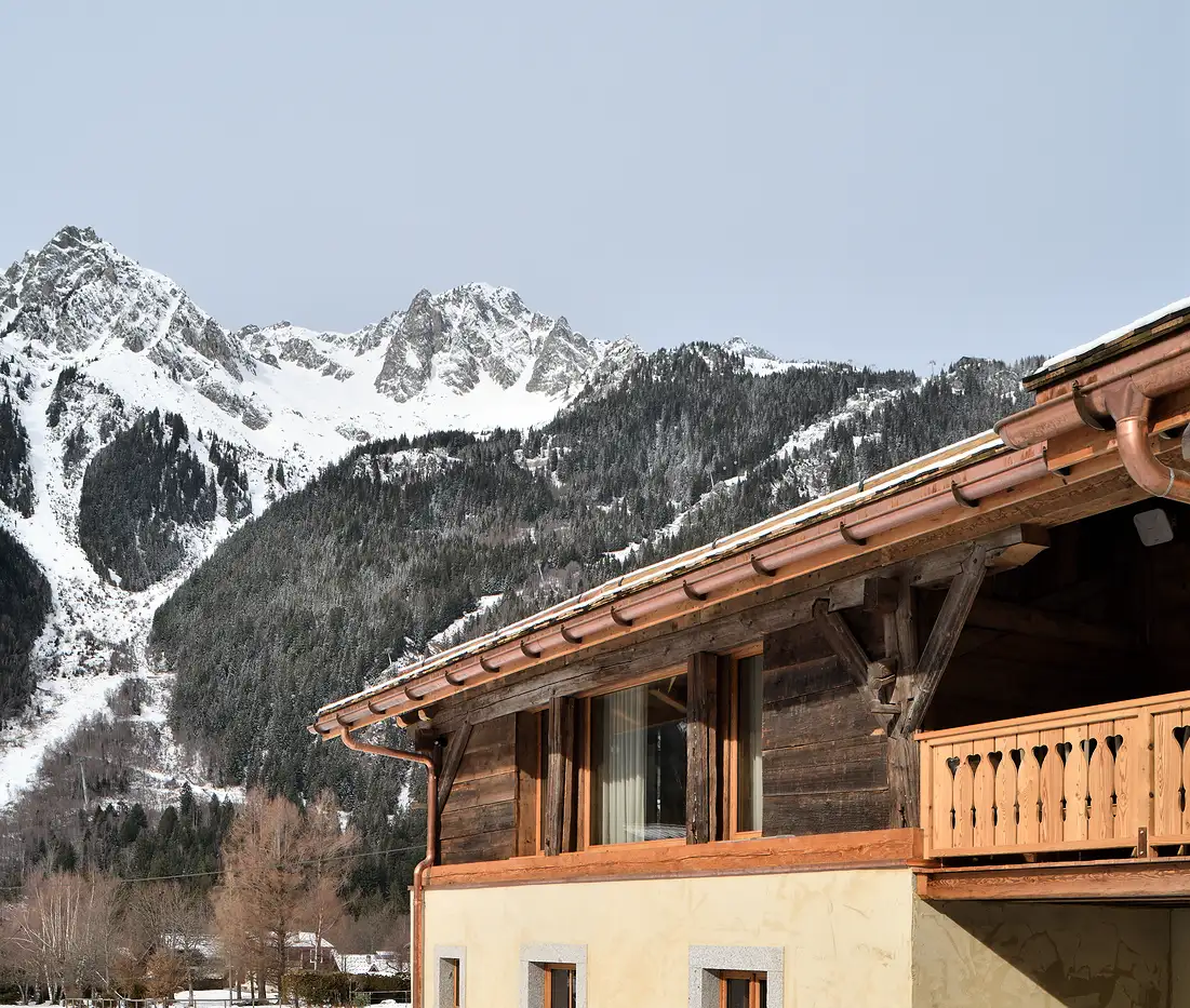 Bâtiment en bois avec balustrade en balcon et montagnes enneigées en arrière-plan, La Ferme des Drus, Chamonix-Mont-Blanc, France.