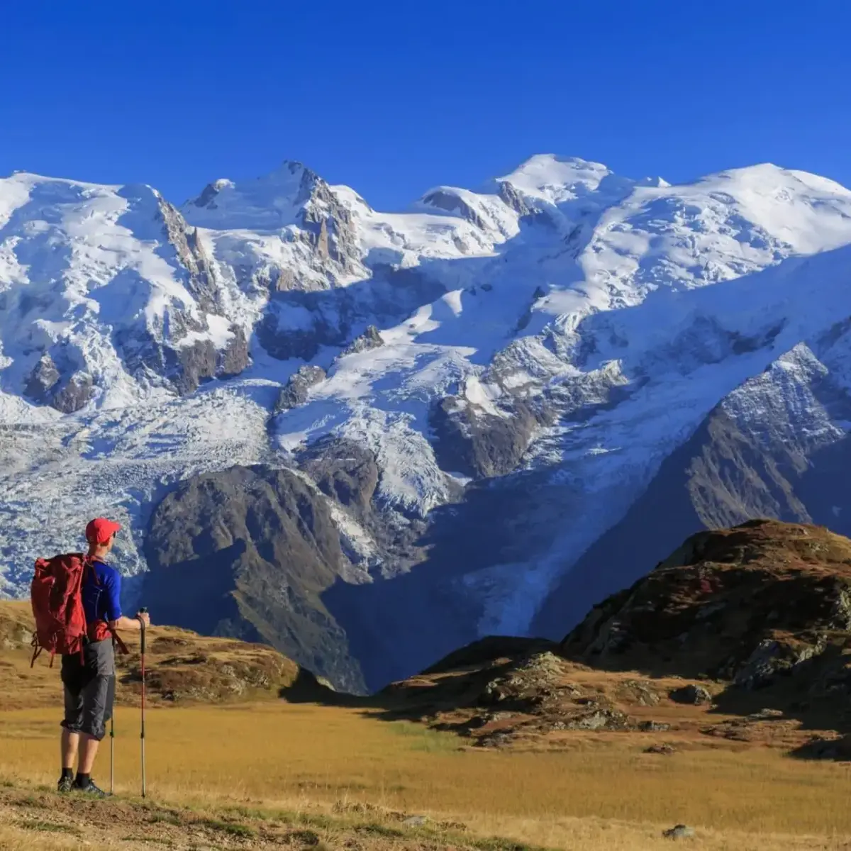 Randonneur avec sac à dos rouge admirant les montagnes enneigées sous un ciel bleu clair, La Ferme des Drus, Chamonix-Mont-Blanc, France.