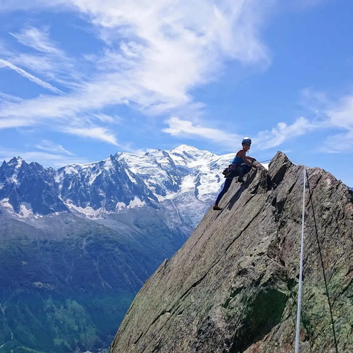 Grimpeur équipé sur une grande paroi rocheuse avec un ciel bleu et montagnes enneigées en arrière-plan, La Ferme des Drus, Chamonix-Mont-Blanc, France.
