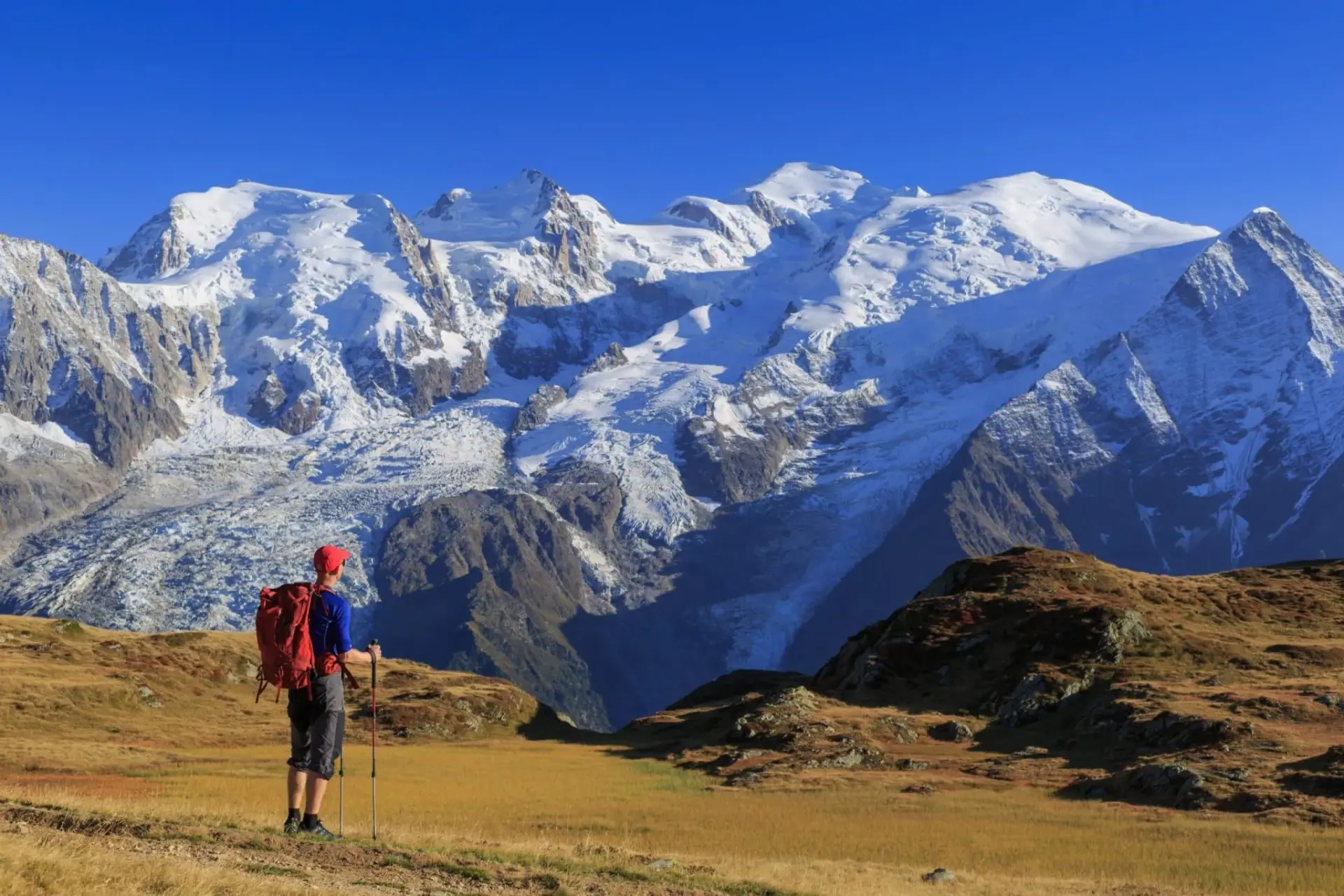 Randonneur avec sac à dos rouge regarde les montagnes enneigées sous un ciel bleu clair, La Ferme des Drus, Chamonix-Mont-Blanc, France.