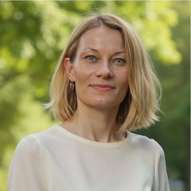 Smiling Justyna Gwardys with shoulder-length hair wearing a white blouse outdoors with green blurred background.